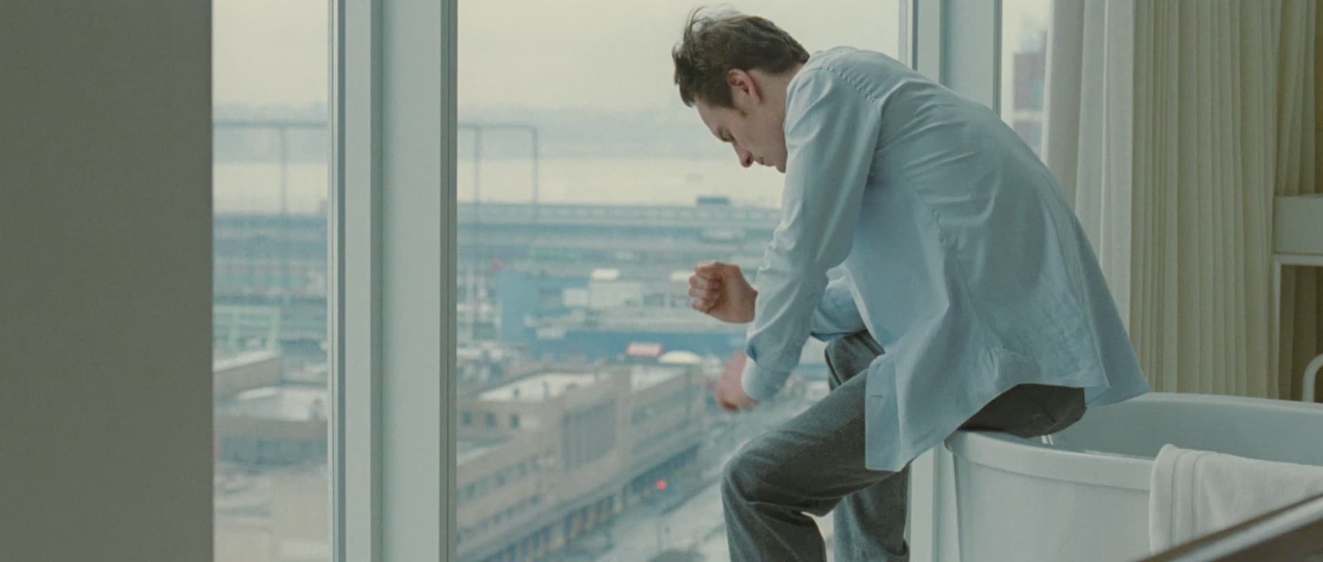 A lonely man sits by a window in an apartment overlookinng New York City